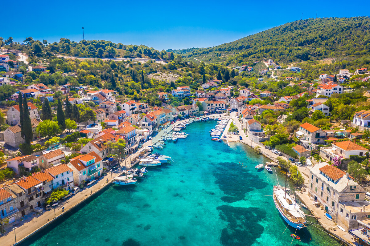 Eine atemberaubende Luftaufnahme von Stomorska auf der Insel Šolta in Kroatien mit dem lebhaften, von Booten gefüllten Yachthafen, der charmanten Uferpromenade und den traditionellen Steinhäusern, alles in idyllische Sommerfarben getaucht.