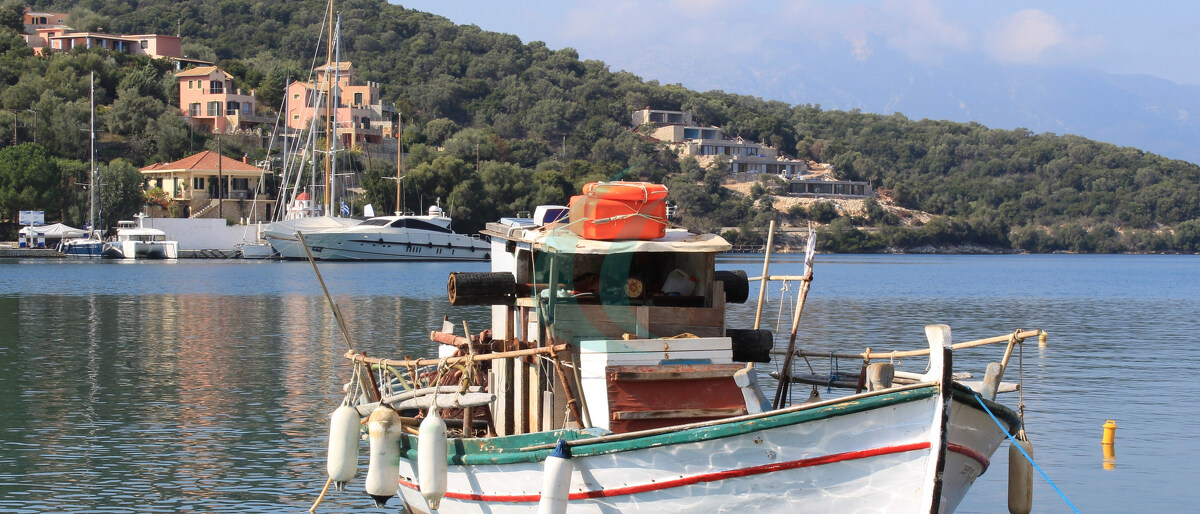 Altes hölzernes Fischerboot im ruhigen blauen Meerwasser am Hafen des Dorfes Vathy auf der Insel Meganisi in Lefkada, Griechenland. Im Hintergrund sind die Yachten und Segelboote des Dorfes zu sehen.