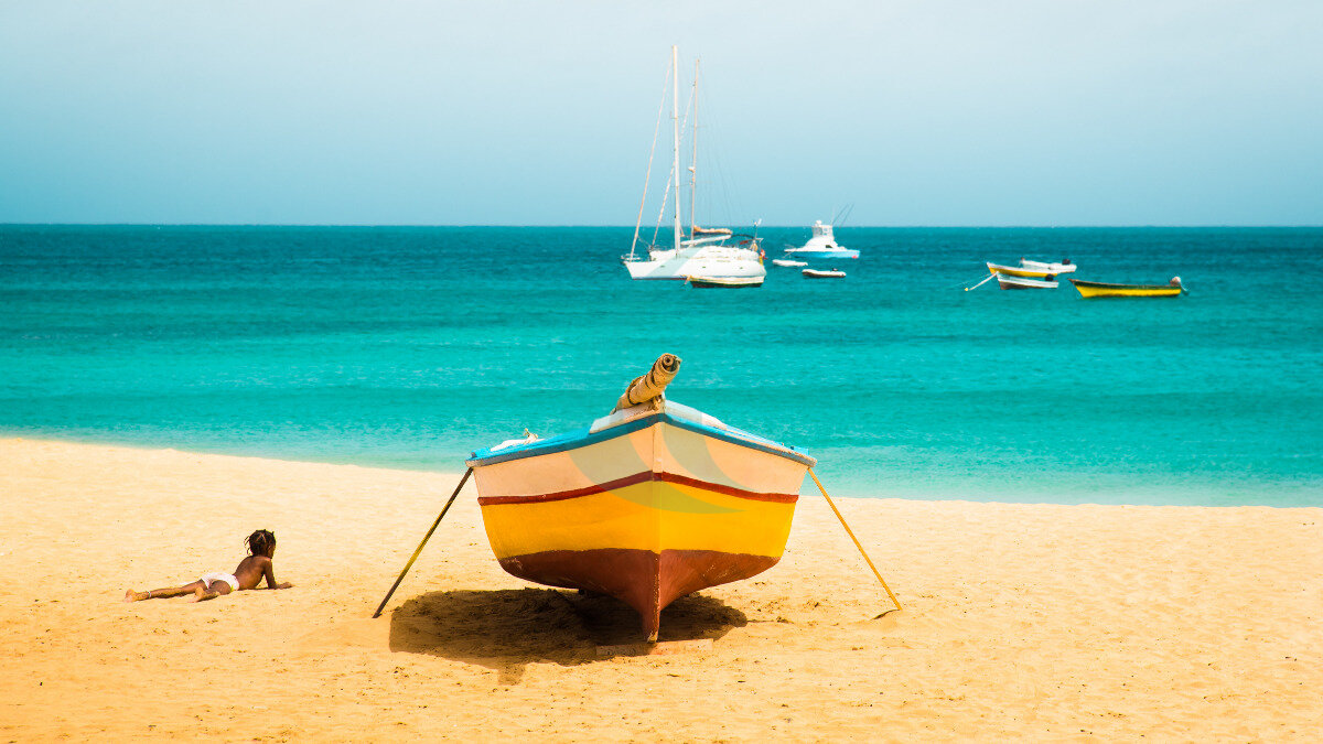 Kapverdische Inseln: Sonniger tropischer Urlaubshintergrund auf der Insel Sal mit der Silhouette eines afrikanischen Kindes am Sandstrand von Santa Maria, bunten Booten und einer türkisfarbenen Meereslandschaft
