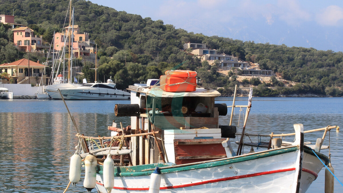 Altes hölzernes Fischerboot in ruhigem blauem Meerwasser im Hafen des Dorfes Vathy auf der Insel Meganisi in Lefkada, Griechenland. Im Hintergrund sind die Yachten und Segelboote des Dorfes zu sehen.