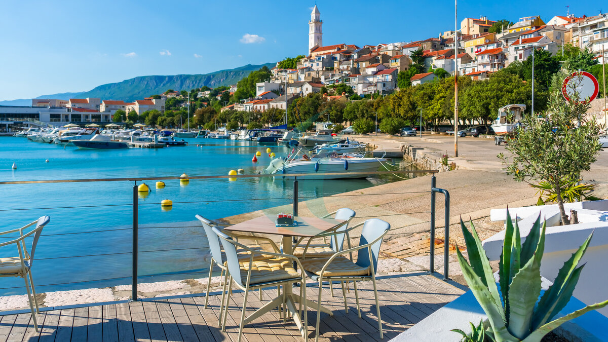 Terrasse eines Restaurants im Freien mit Blick auf die Adria und die Uferpromenade von Novi Vinodolski an einem sonnigen Sommertag. Mediterrane Urlaubsatmosphäre