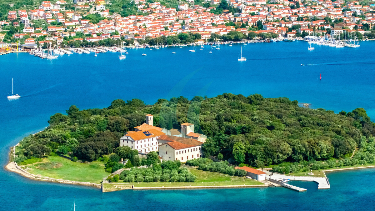 Eine weite Luftaufnahme  - Marina Punat & der Insel Košljun mit ihrem Kloster, umgeben von ruhigem blauem Wasser und der Küstenstadt Punat im Hintergrund. Segelboote verteilen sich über das Meer.