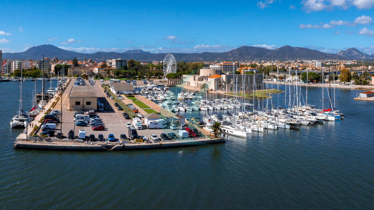 Der lebhafte Yachthafen in Olbia auf Sardinien zeigt Yachten und Boote, ein Riesenrad sowie eine Mischung aus moderner und traditioneller Architektur vor sanft geschwungenen Hügeln.